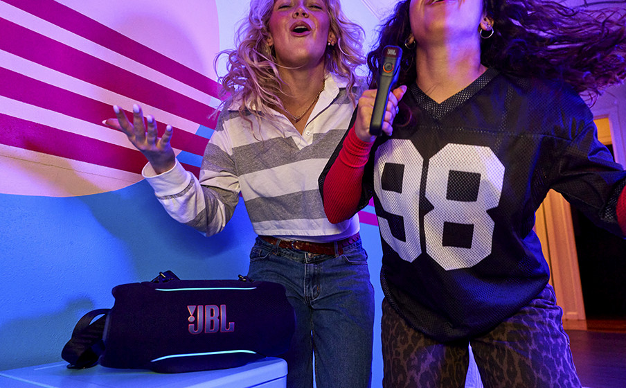 Zwei Frauen singen in einen JBL-Lautsprecher mit bunten Lichtern und gestreiftem Hintergrund.