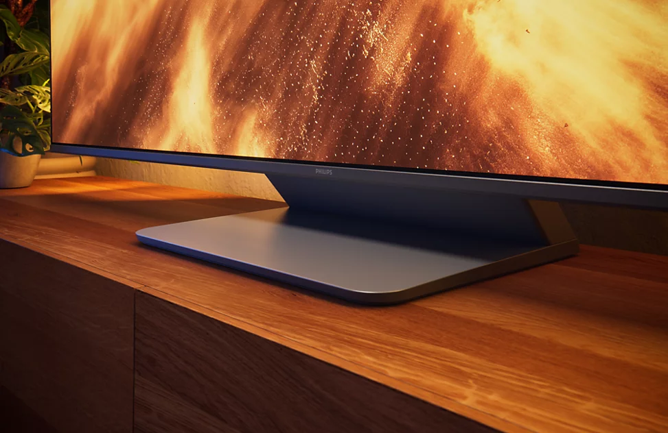 Ein moderner Fernseher mit silbernem Standfuß steht auf einem Holzmöbel. Im Hintergrund ist ein flammendes Bild zu sehen.