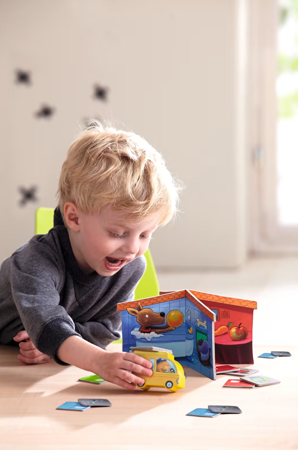 Egy kisfiú sárga játékautóval és egy játékházzal játszik az asztalon.