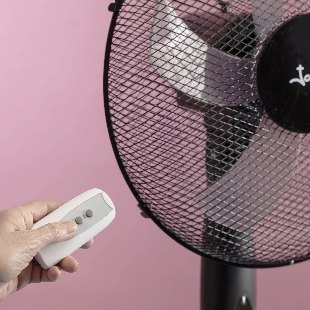 Una mano sostiene un mando a distancia blanco frente a un ventilador sobre fondo rosa.