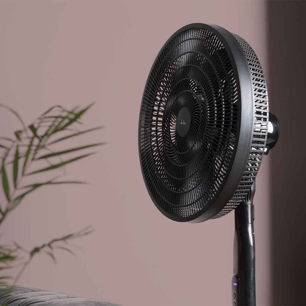 Un ventilador de pie negro está frente a una pared rosa, con una rama de planta a la izquierda.