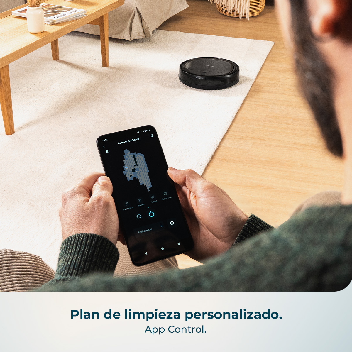 Una persona sostiene un smartphone con una app de limpieza y un mapa. Un robot aspirador está en una alfombra.