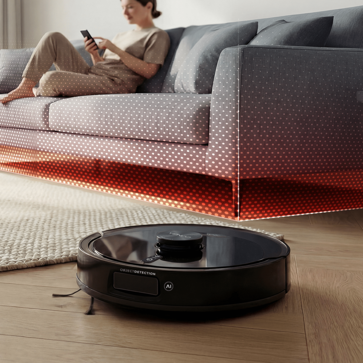 Un robot aspirador limpia el suelo bajo un sofá donde se sienta una persona.