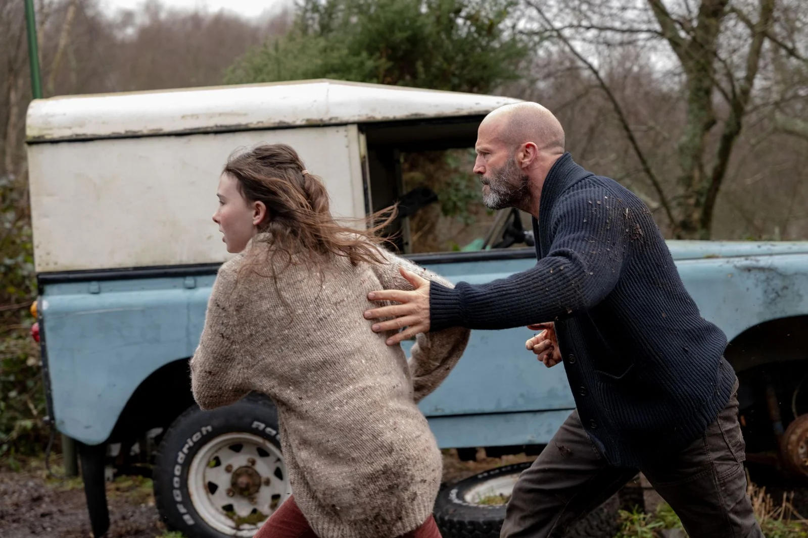 Mann mit Glatze und Frau rennen vor einem alten Geländewagen.