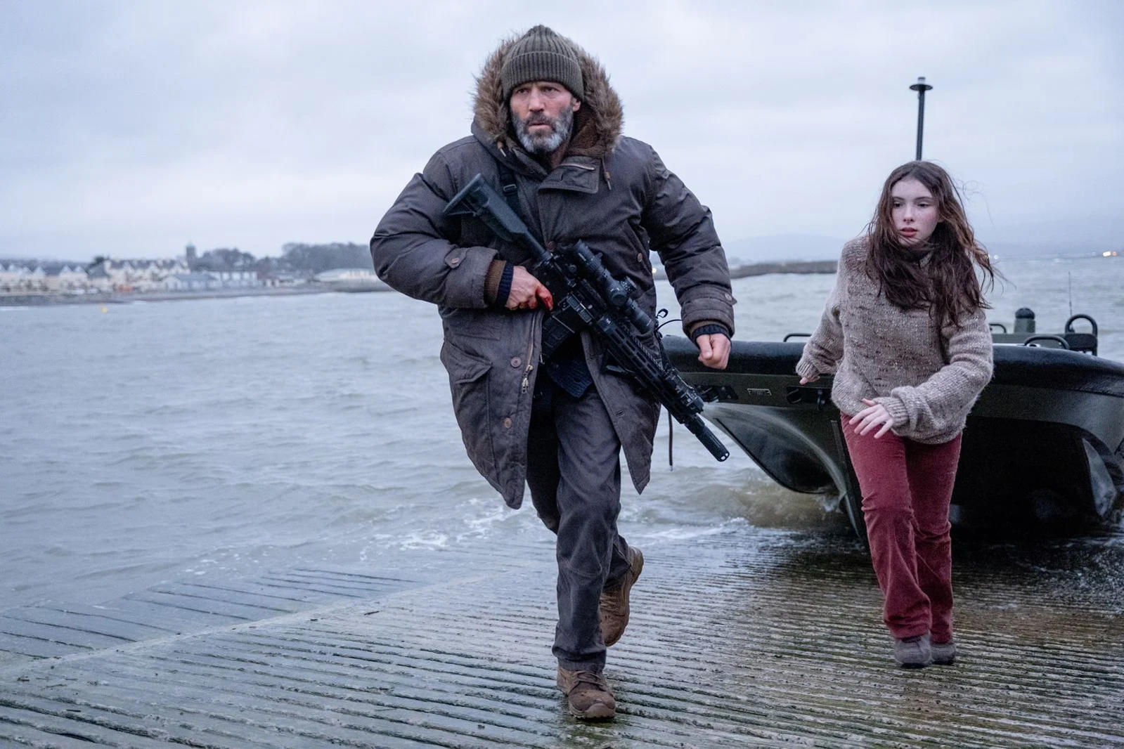 Ein Mann mit Gewehr und ein Mädchen laufen auf einer nassen Rampe neben einem Boot.