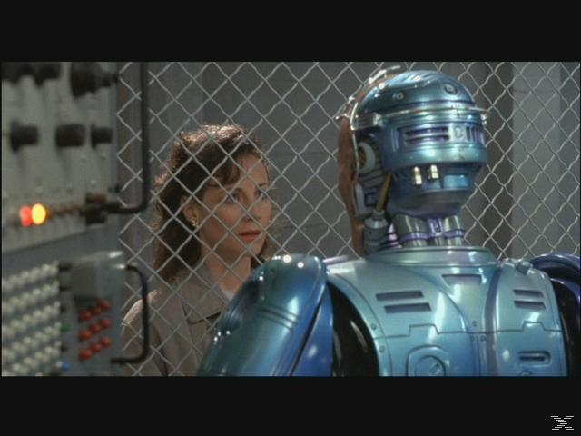 Eine Frau blickt durch einen Zaun auf einen blauen Roboter.