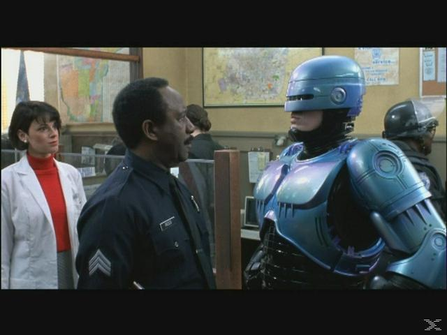 Ein Polizist spricht mit RoboCop. Eine Frau steht im Hintergrund.