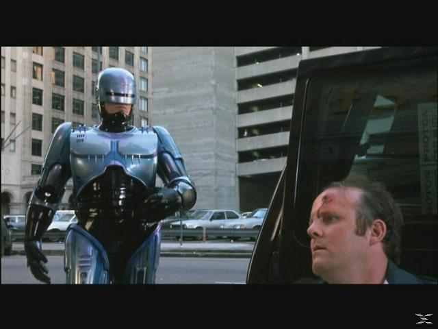 RoboCop steht neben einem verletzten Mann, der aus einem Auto steigt.