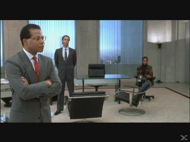Drei Geschäftsleute in einem Büro. Ein Mann im Vordergrund mit verschränkten Armen.