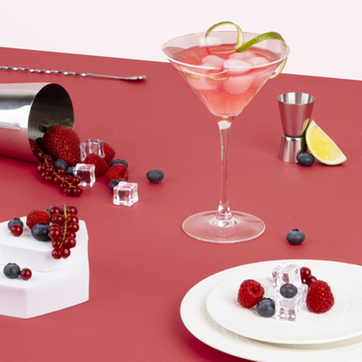 Un cocktail rosa con ghiaccio e bacche, accanto a uno shaker e frutta.