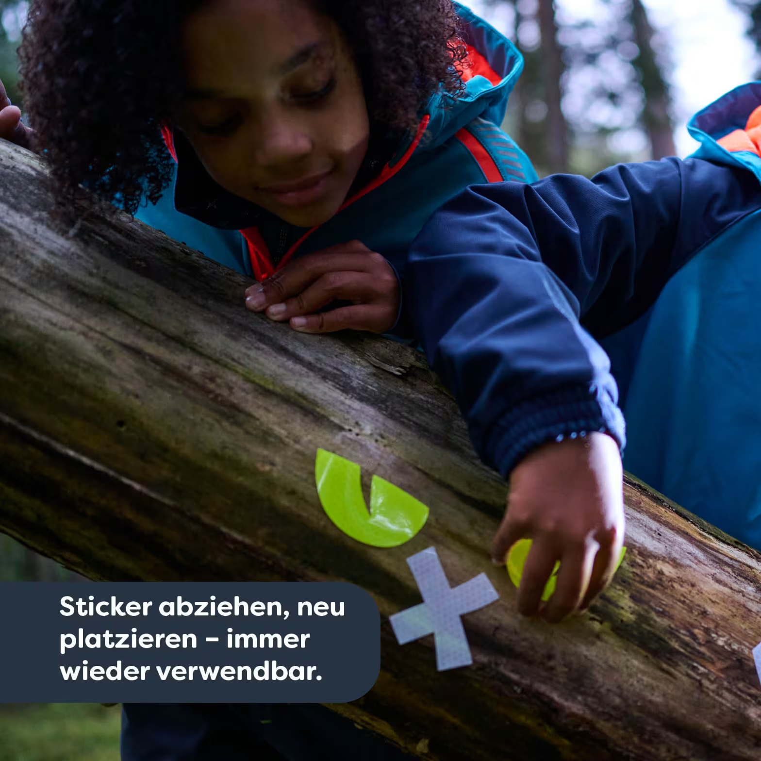 Egy gyerek az erdőben fatörzsön mászik, élénksárga matricákkal játszik.
