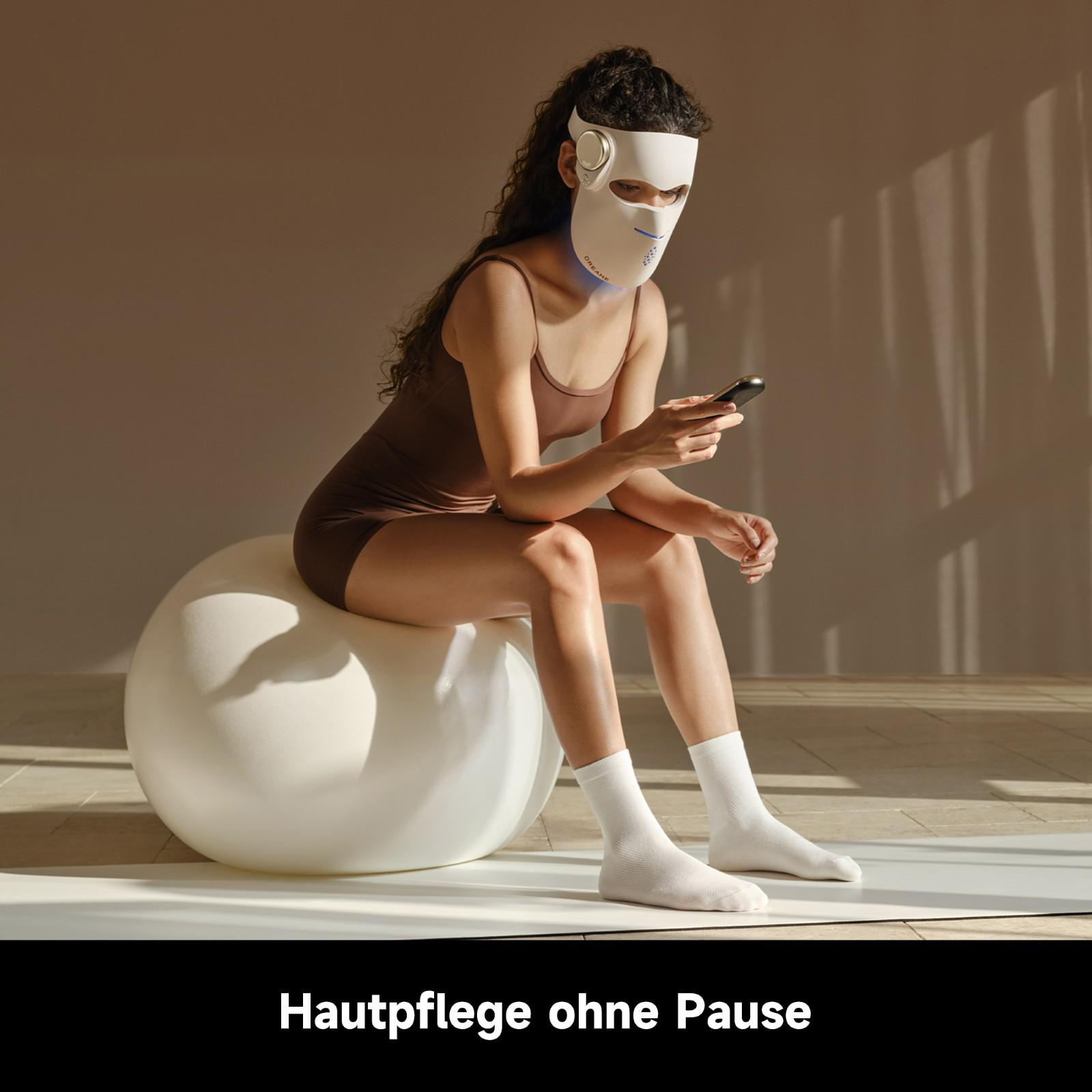 Frau sitzt auf Ball, trägt LED-Maske und hält Smartphone.