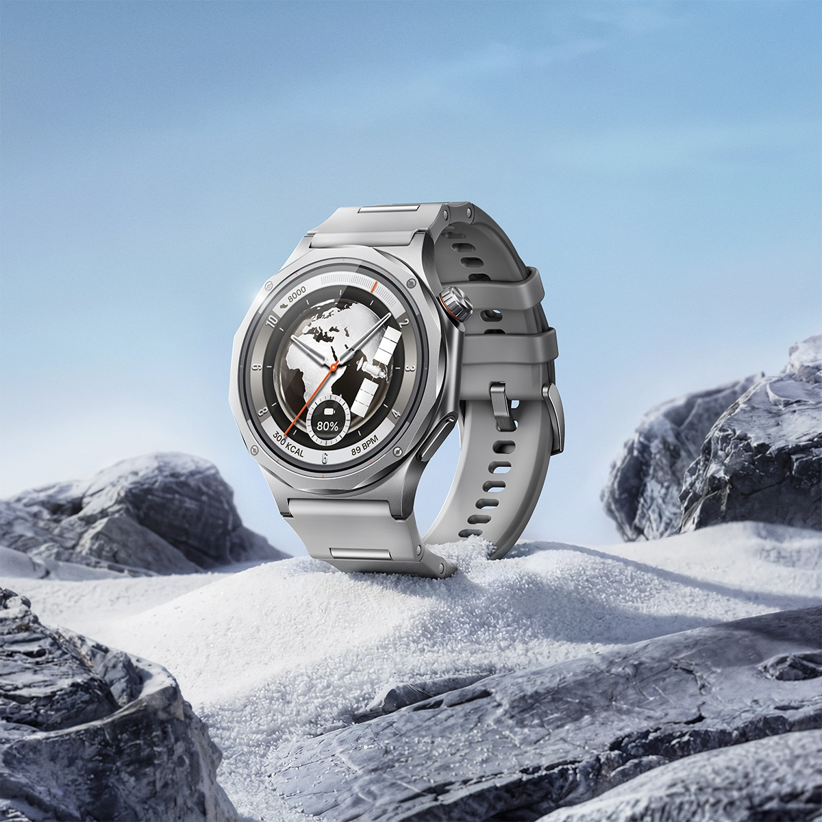 Reloj inteligente gris sobre nieve con rocas y cielo azul.