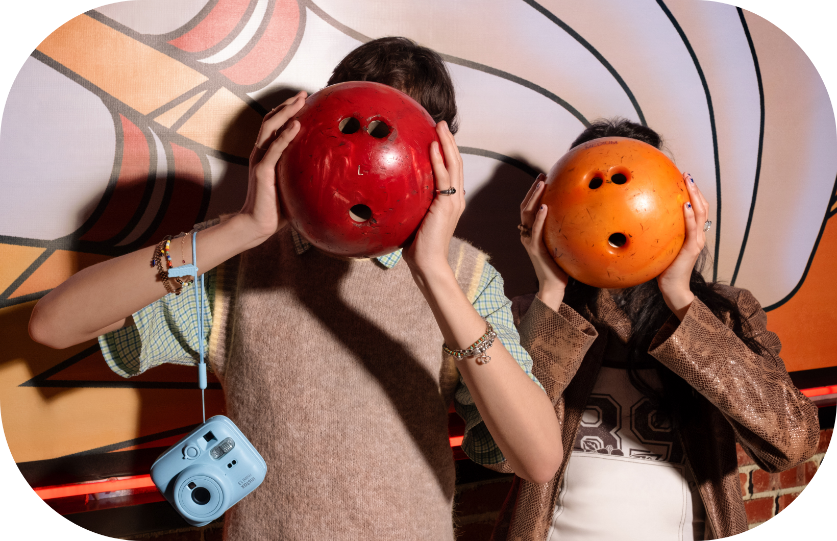 Zwei Personen halten Bowlingkugeln vor ihre Gesichter. Links hängt eine blaue Kamera.