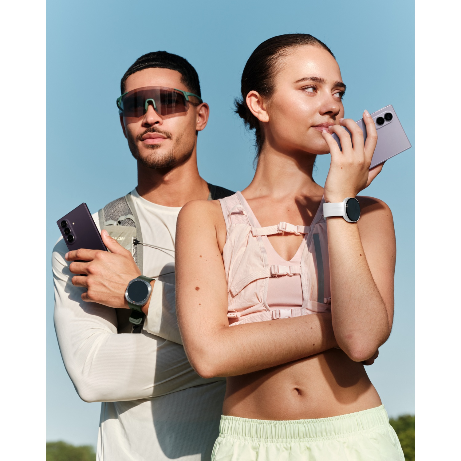 Man and woman with phones and watches outdoors under a blue sky.