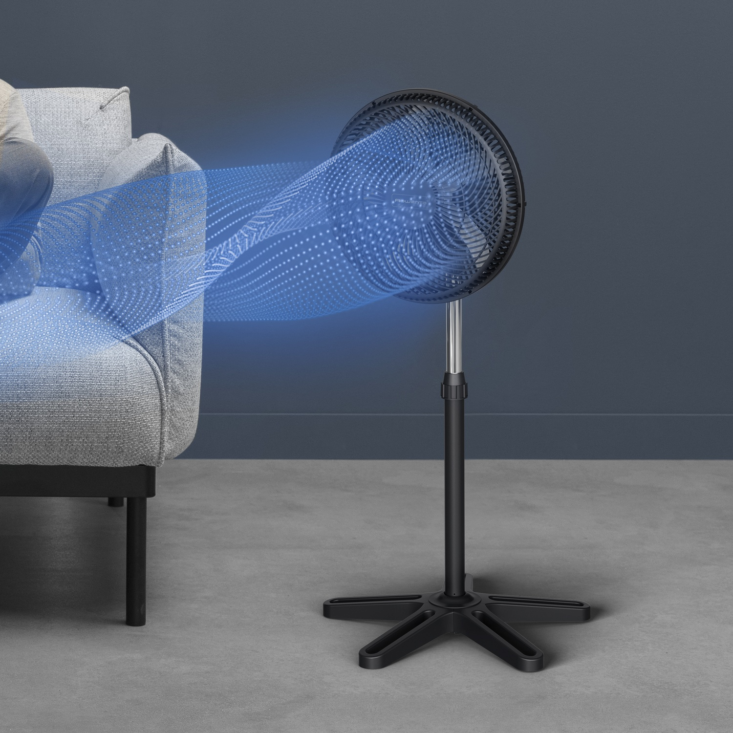A black oscillating fan blows blue air toward a gray sofa on a concrete floor.
