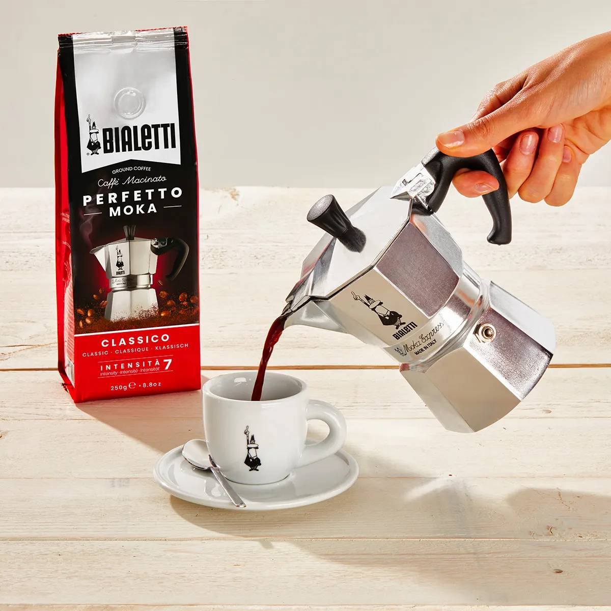 Cafetera Bialetti Moka vertiendo café en una taza, junto a un paquete de café.
