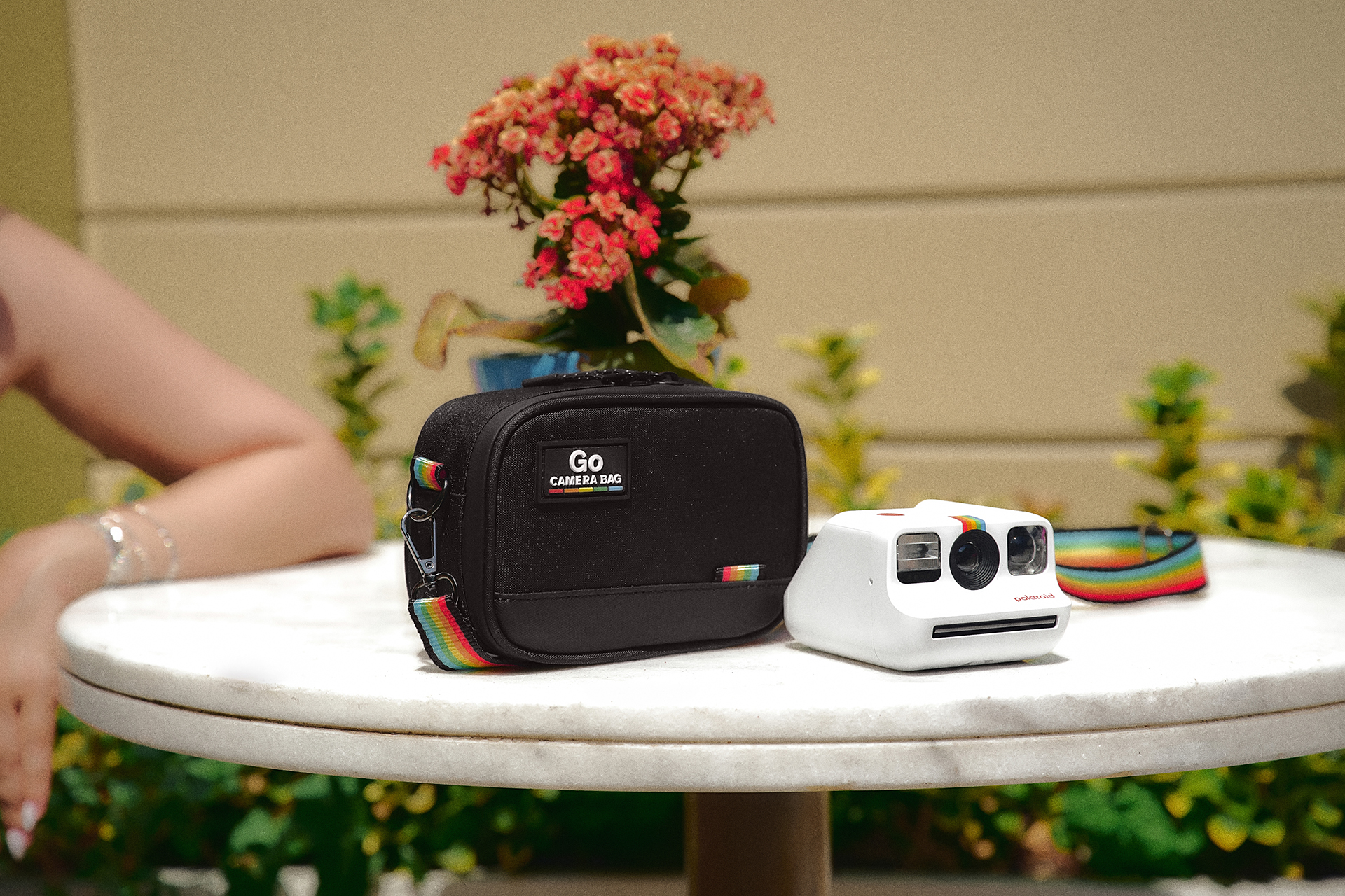 Gökkuşağı detaylı beyaz bir Polaroid kamera ve siyah kamera çantası masada duruyor.