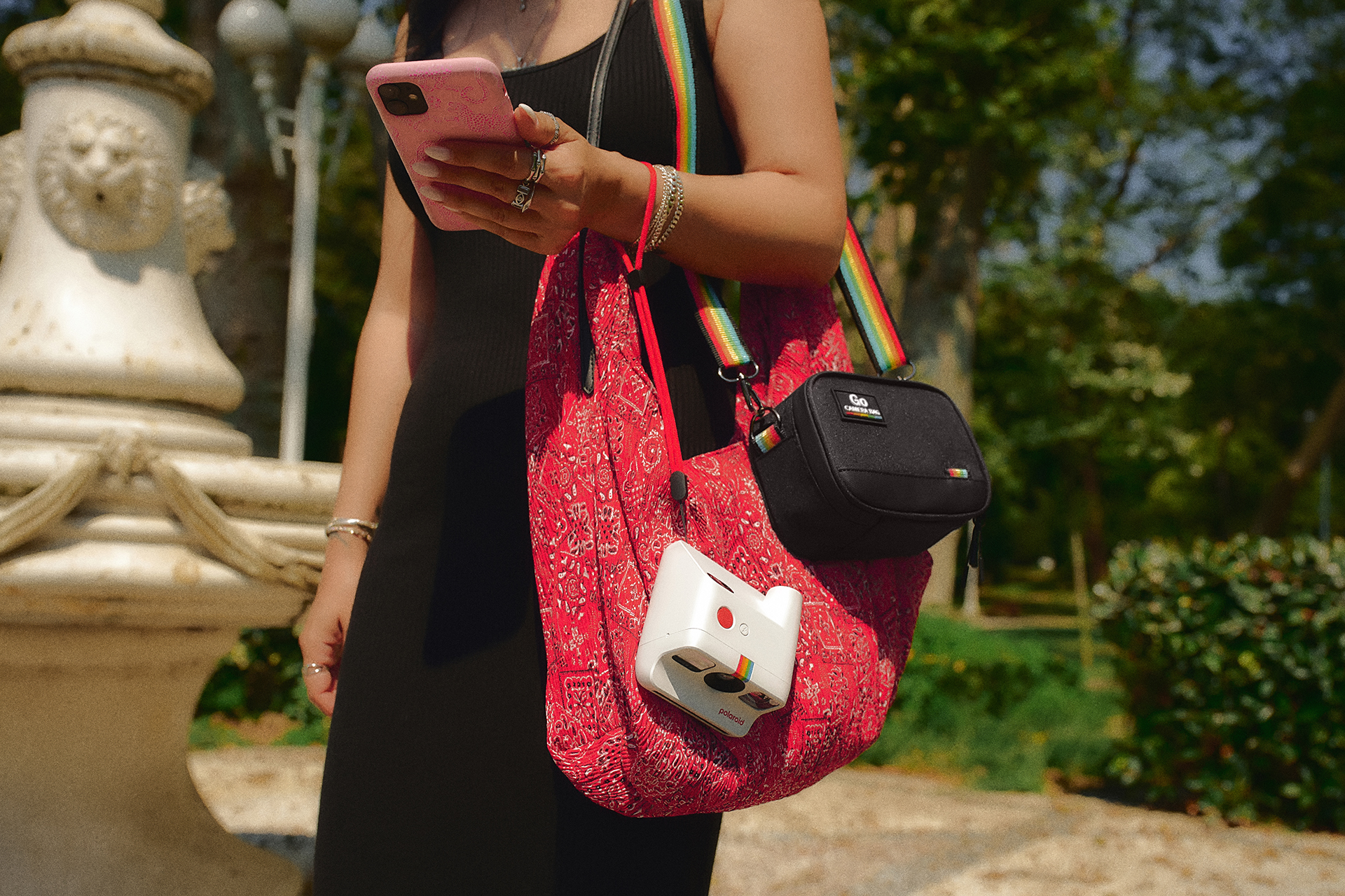 Bir kadın pembe akıllı telefon tutuyor, yanında Polaroid kameralı kırmızı çanta var.