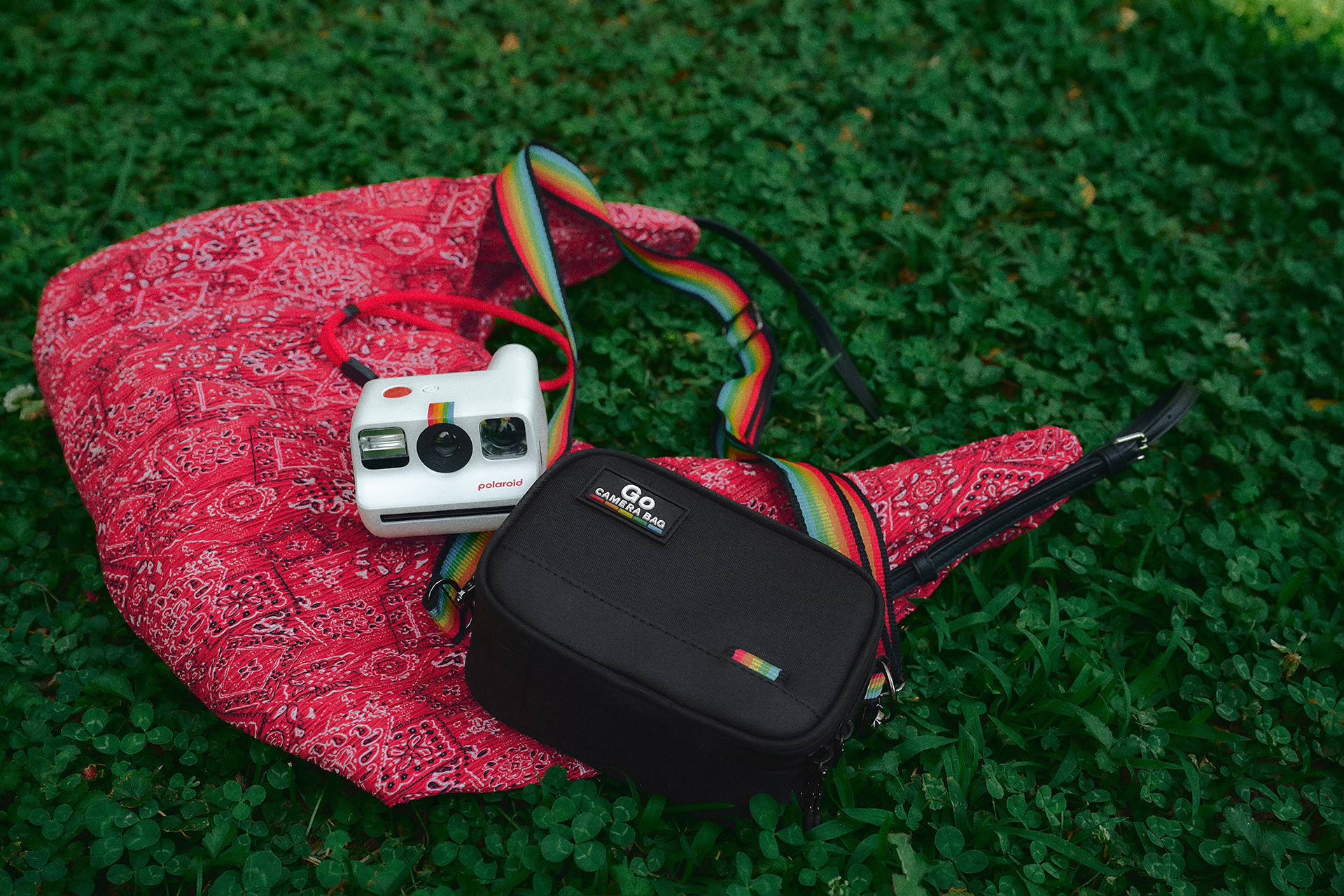 Beyaz bir Polaroid kamera ve siyah bir kamera çantası, çimlerin üzerinde kırmızı bandana kumaşın üzerinde duruyor.