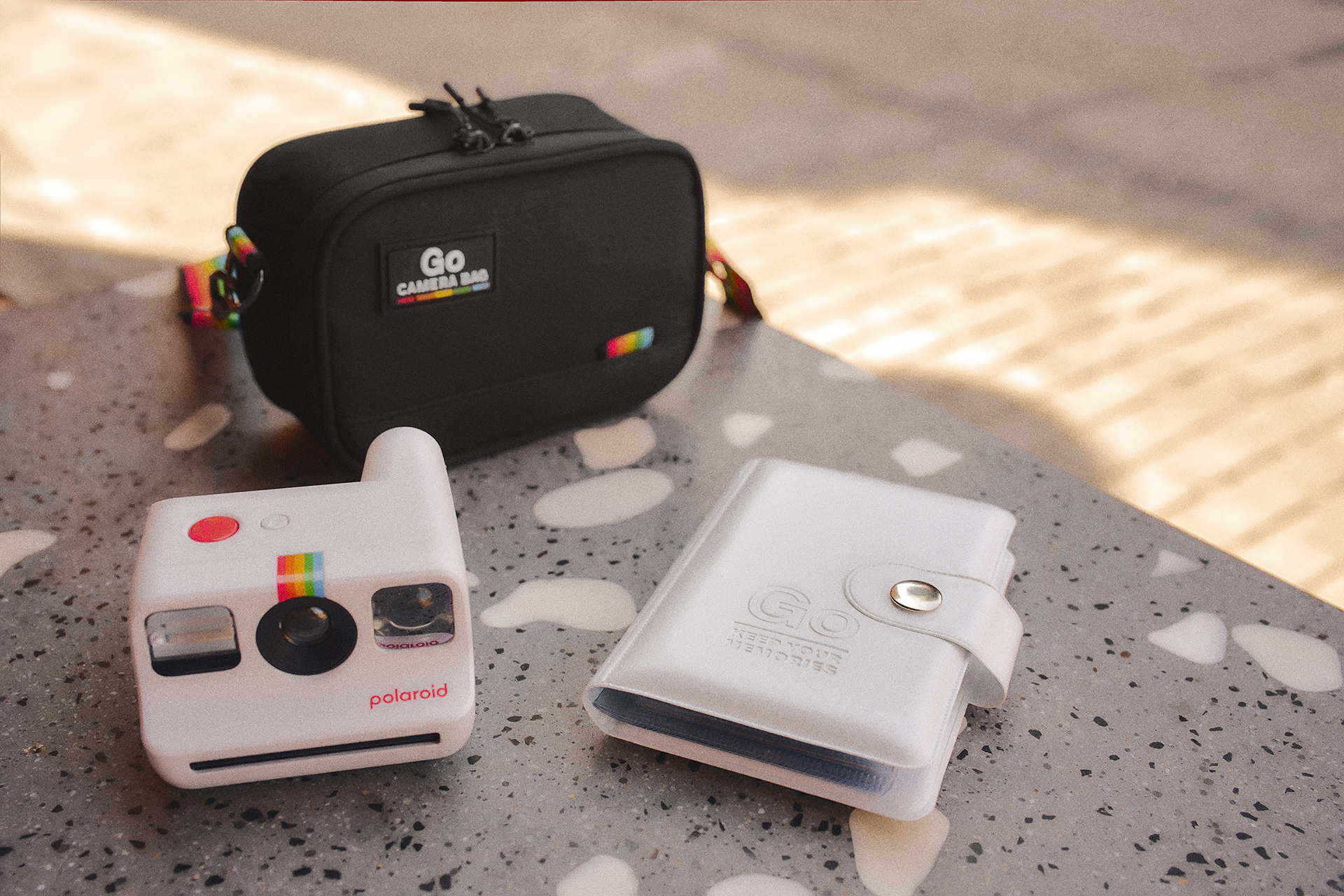 Beyaz Polaroid kamera, siyah kamera çantası ve beyaz fotoğraf albümü masada.