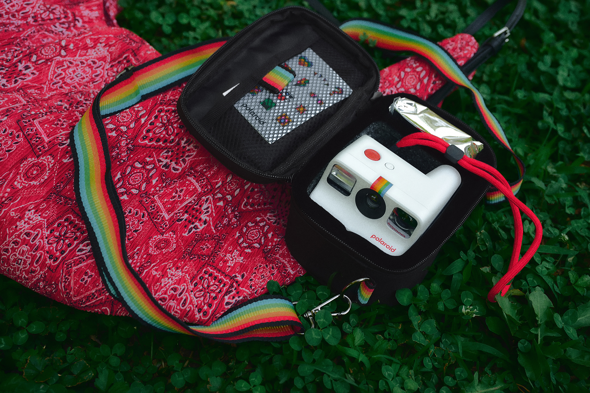Beyaz bir Polaroid kamera, çimenlerin üzerinde siyah bir çantanın yanında kırmızı bandana kumaşın üzerinde duruyor.