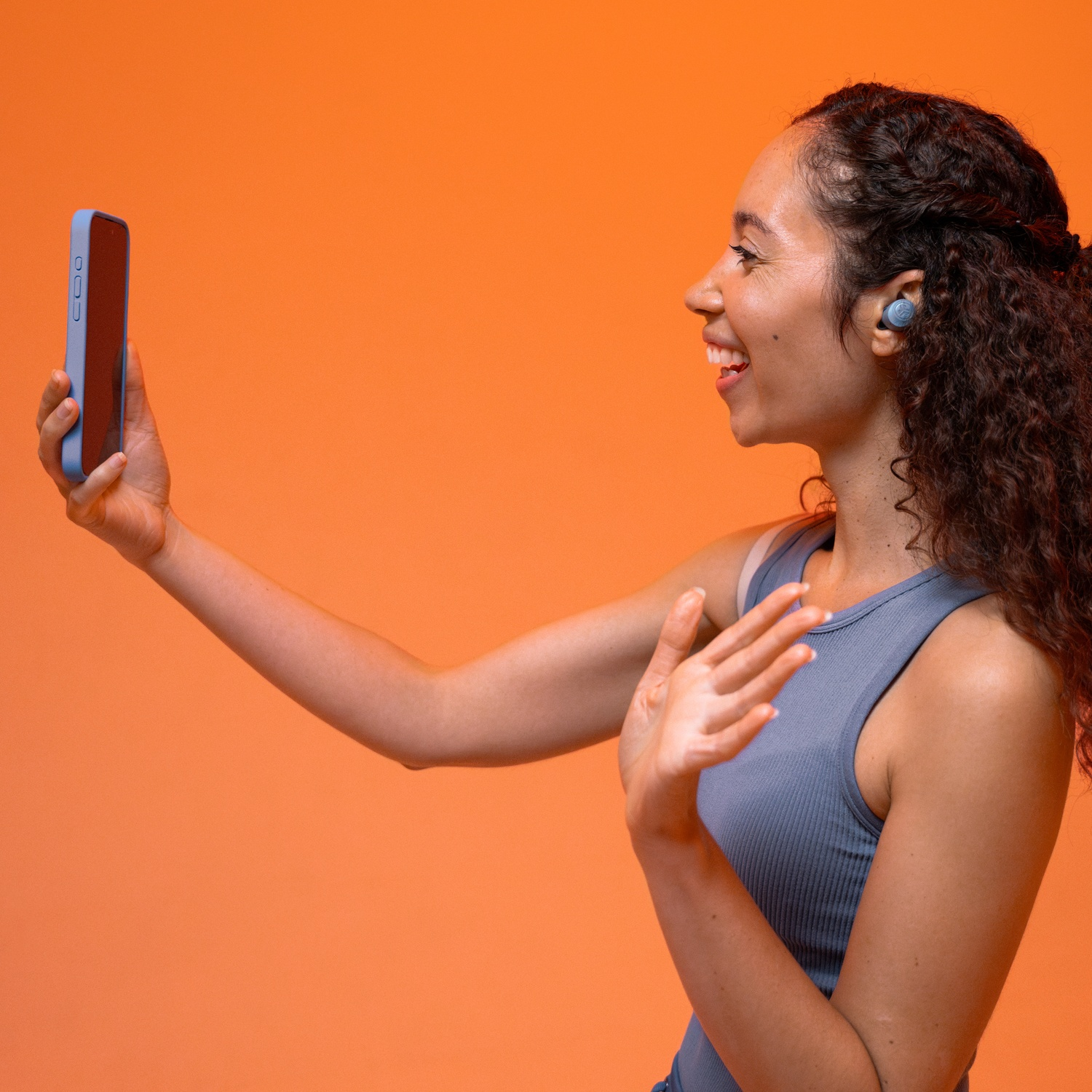 Une femme aux cheveux bouclés tient un smartphone bleu et fait signe.