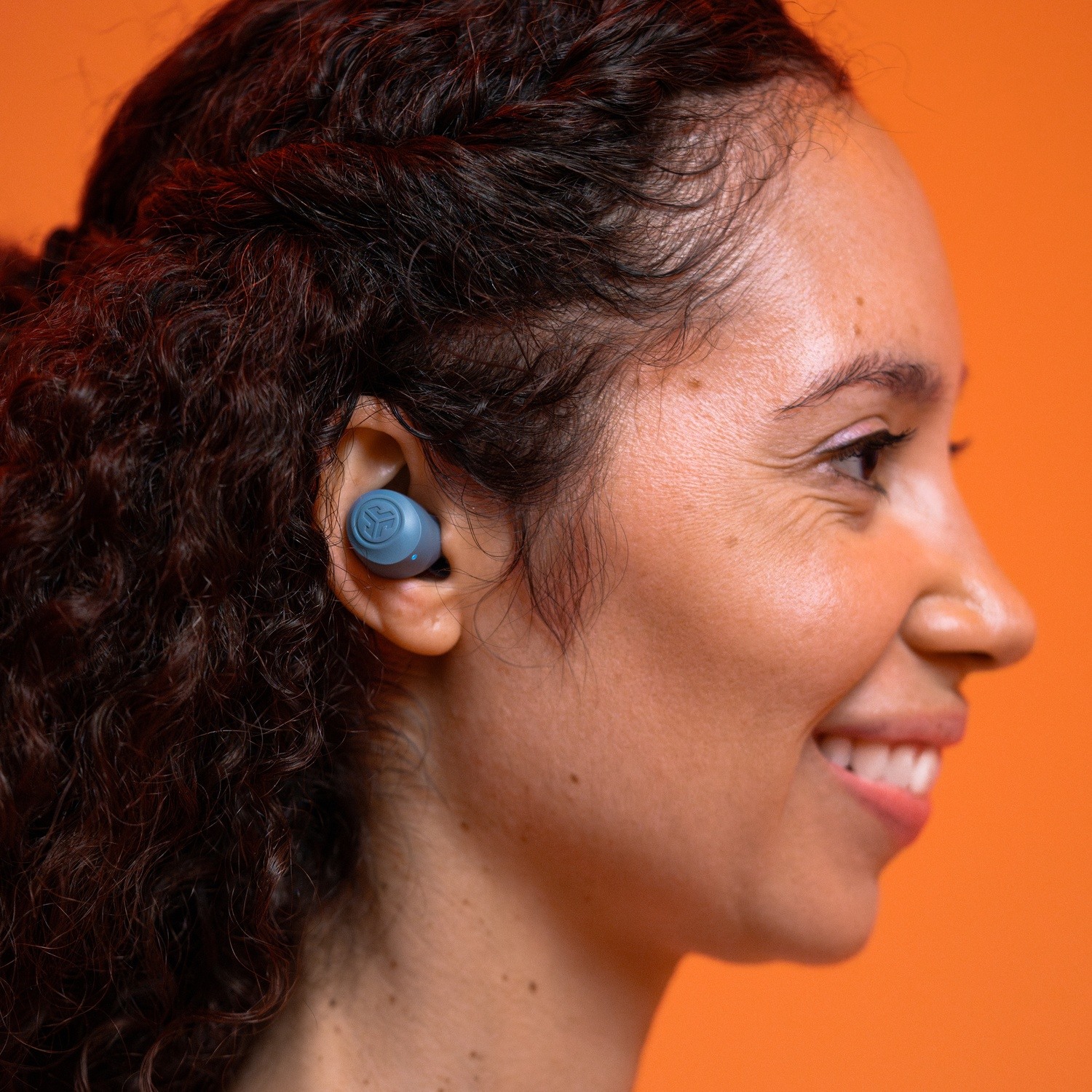 Femme avec des écouteurs bleus dans l'oreille, souriant, sur fond orange.