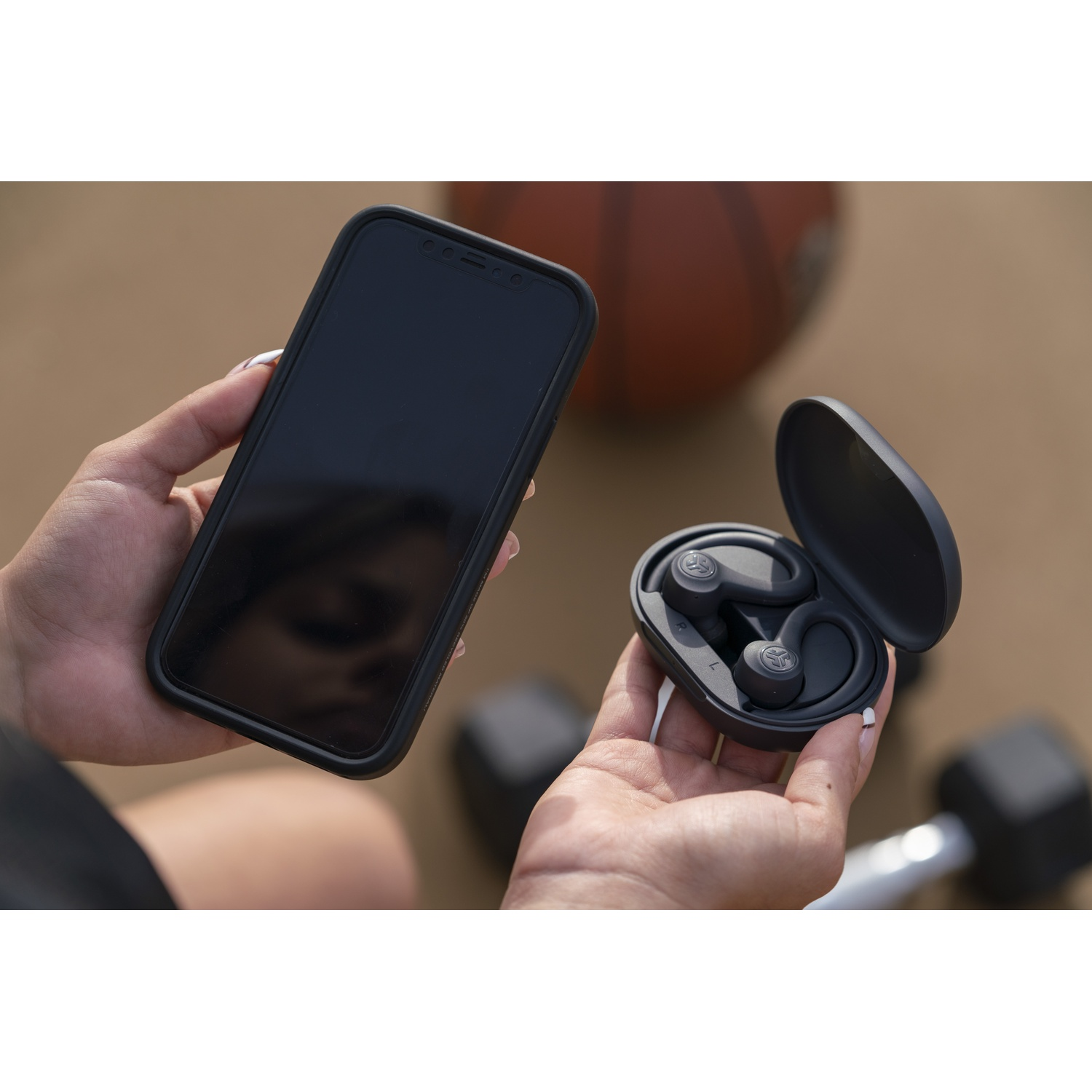 Hands hold a smartphone and open earbuds with charging case. Dumbbells in background.
