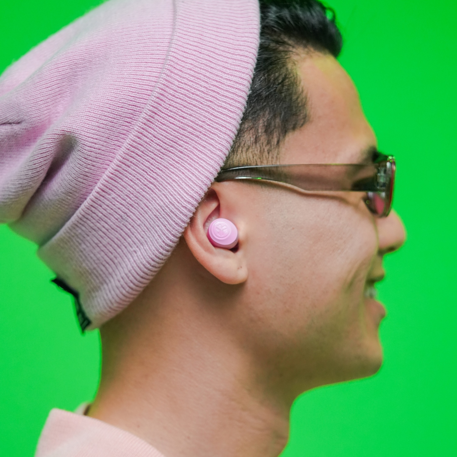 Homme portant un bonnet rose, des lunettes et des écouteurs roses sur fond vert.