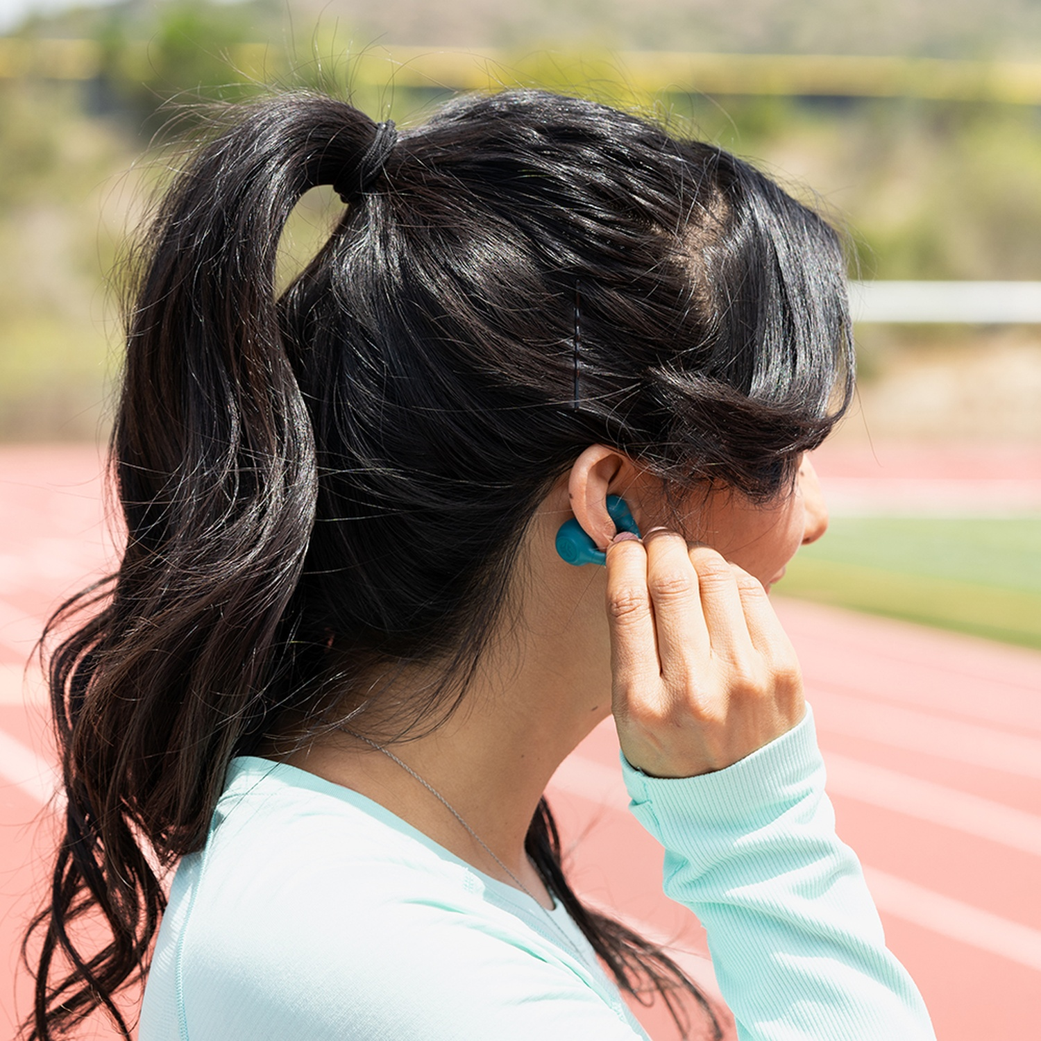 Woman with ponytail inserts a teal earbud into her ear.