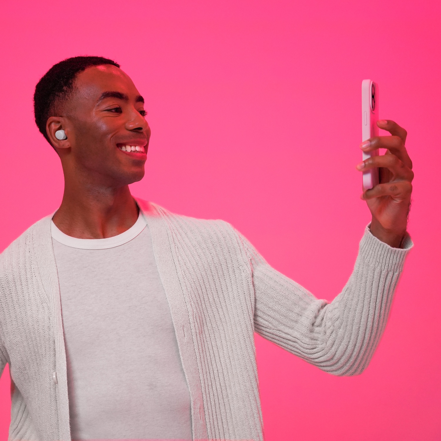 A man wearing earbuds holds a cell phone against a pink background.