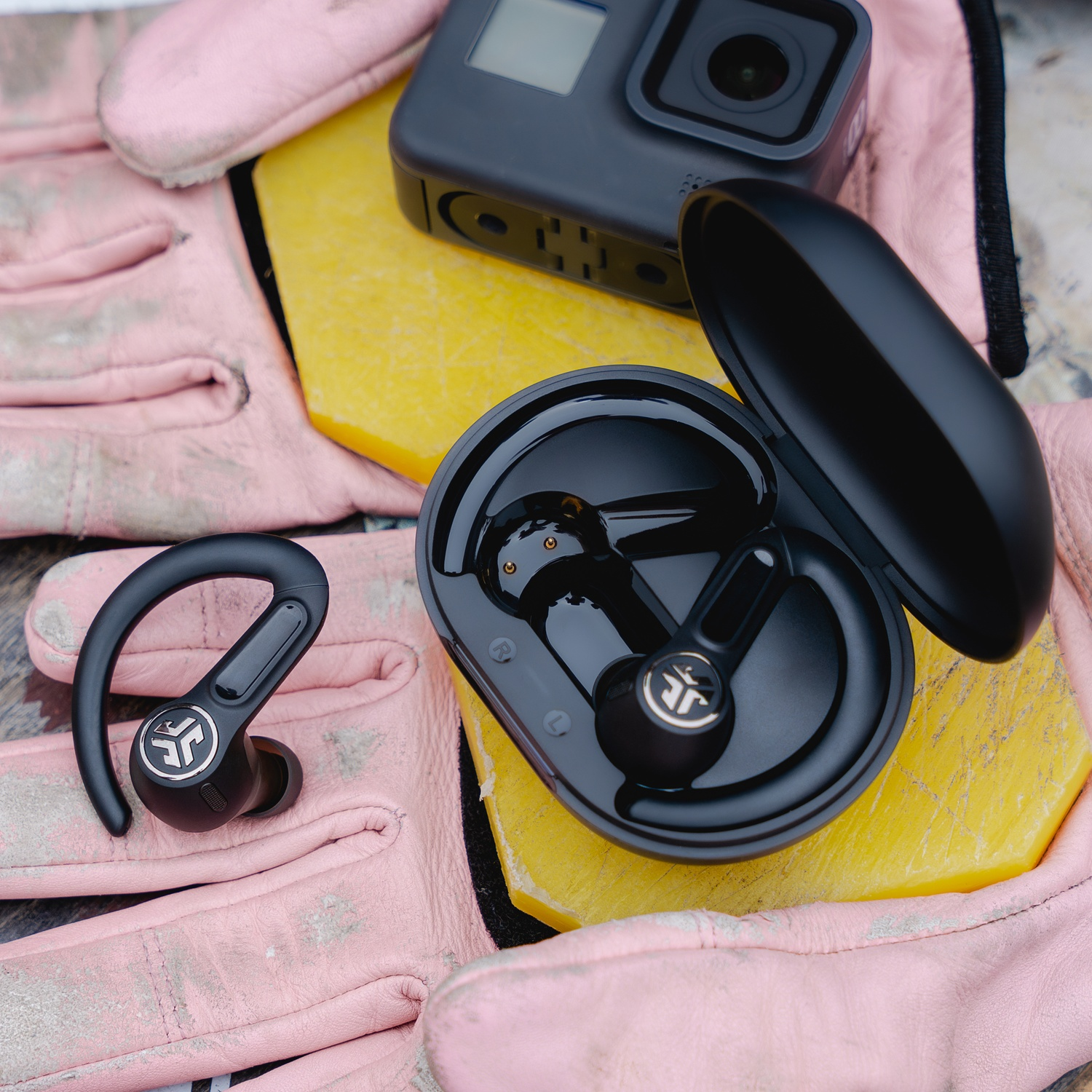 Black earbuds and charging case on pink gloves beside a GoPro camera.