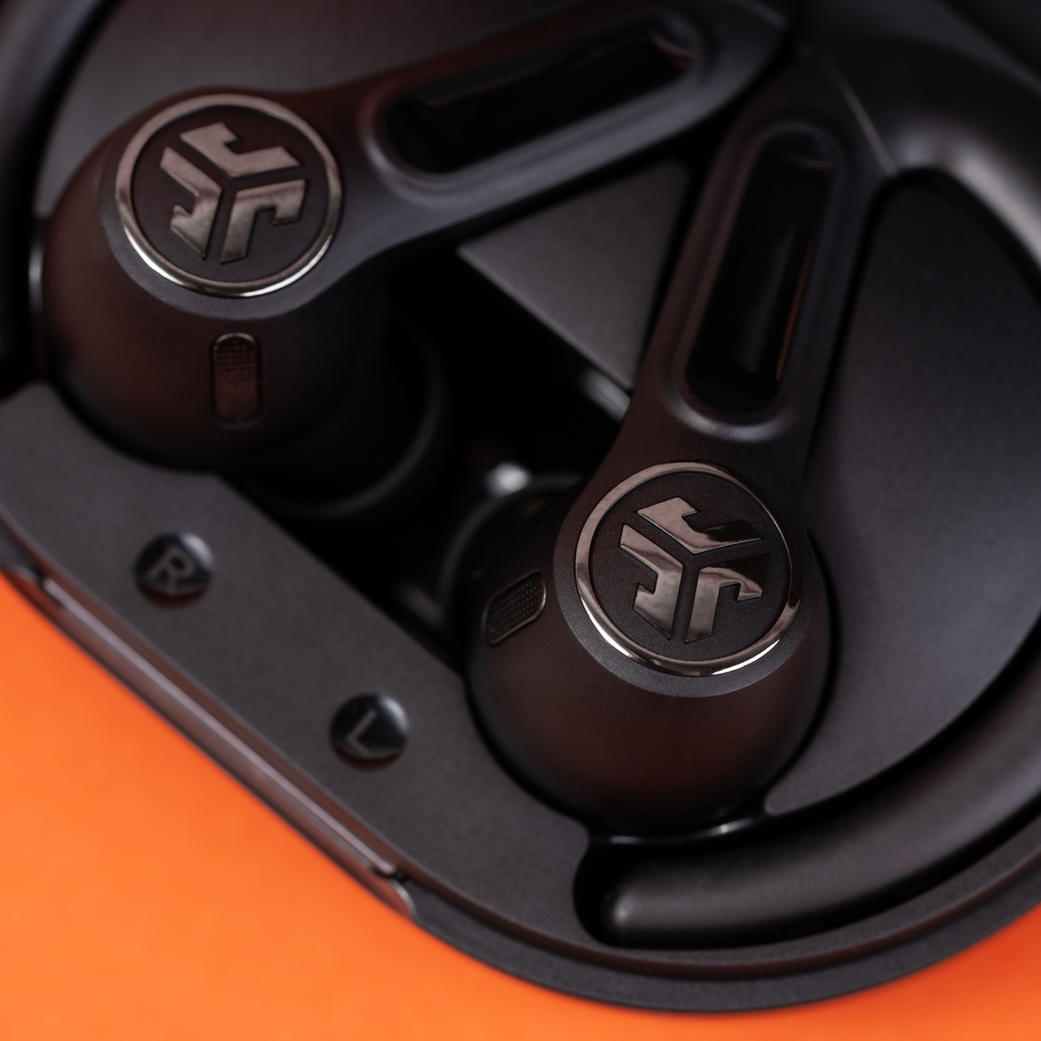 Black wireless earbuds with silver logo in charging case on orange background.
