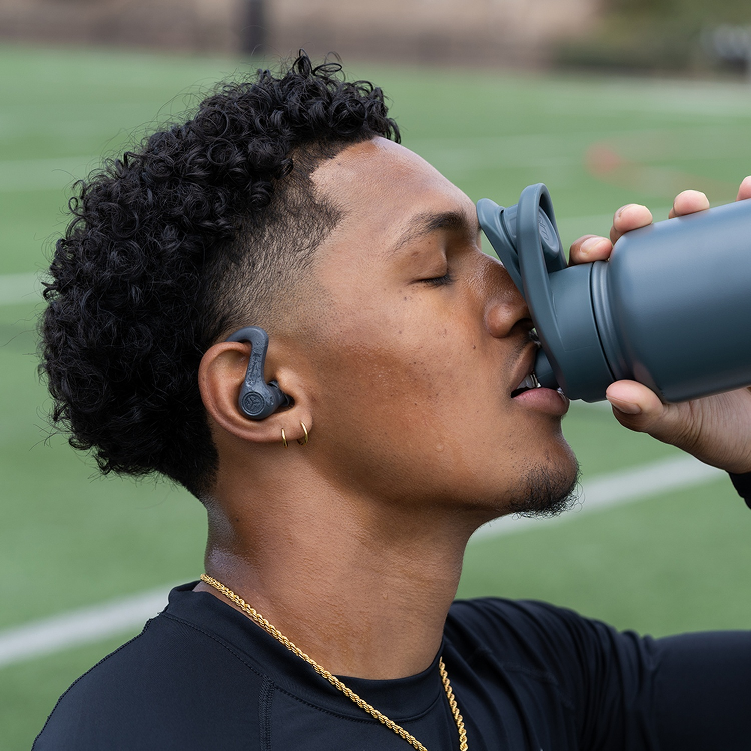 A man with curly hair drinks from a gray water bottle on a green field.