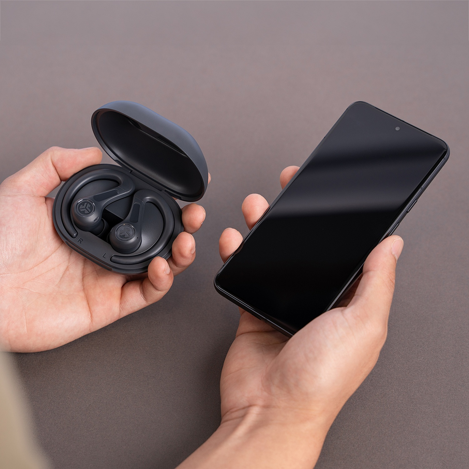Hands hold black earbuds in case and a smartphone on a brown background.