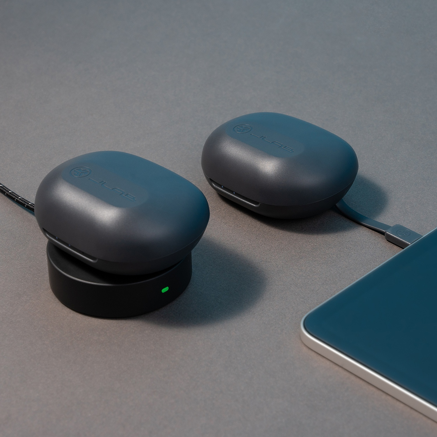 Two gray JLab earbud cases. One on a black charging base, one connected to a phone.
