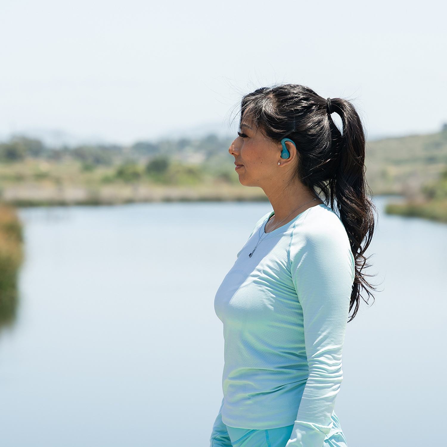 A woman with a ponytail and blue earpiece looks out over a lake.