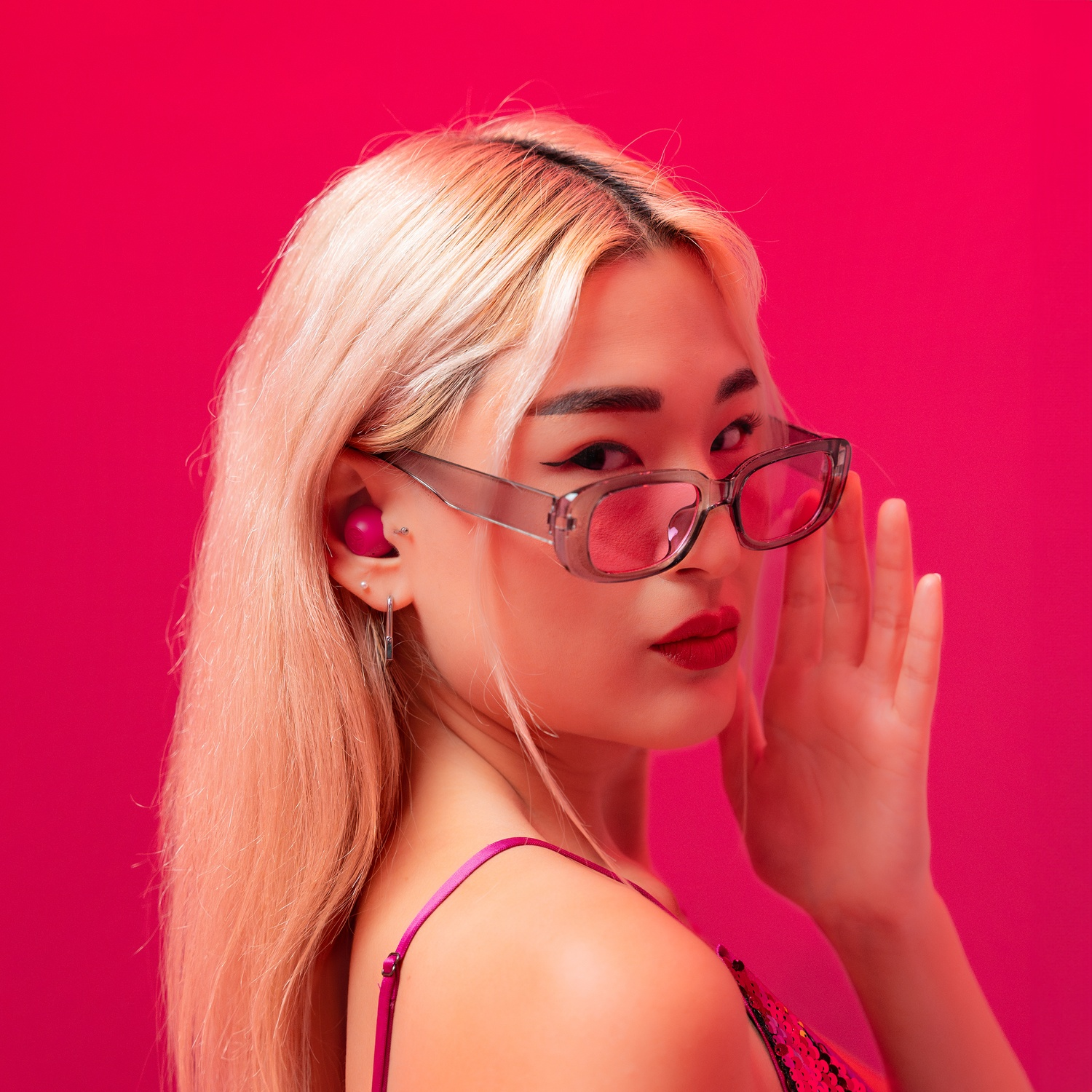 Een vrouw met blond haar en zonnebril draait zich om, tegen een roze achtergrond.