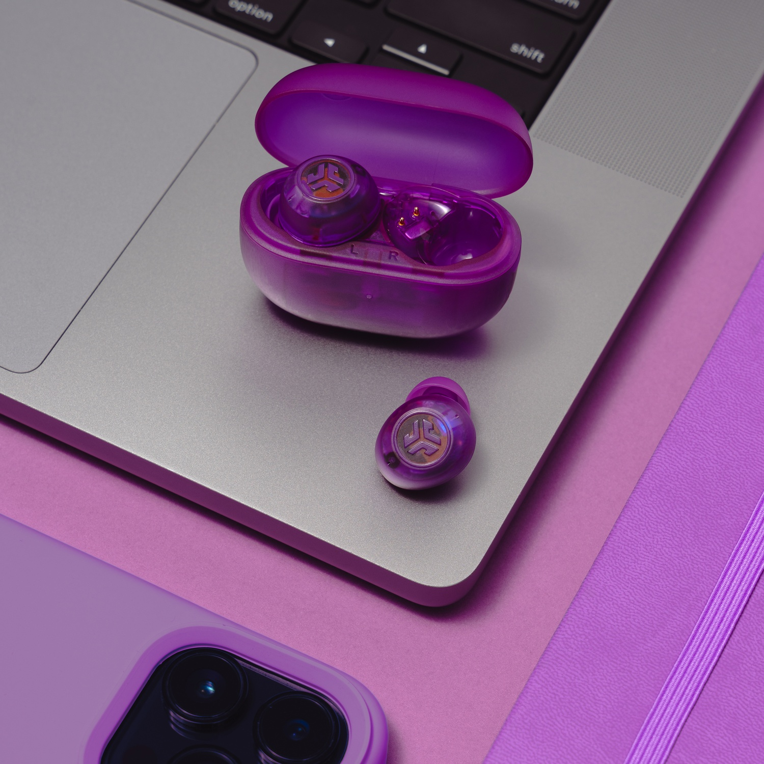 Paarse draadloze oordopjes in hoesje op laptop, naast telefoon en notitieboek.
