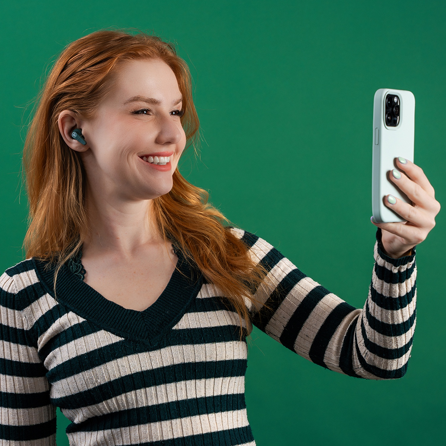 Femme rousse en pull rayé prend un selfie avec téléphone et écouteur.