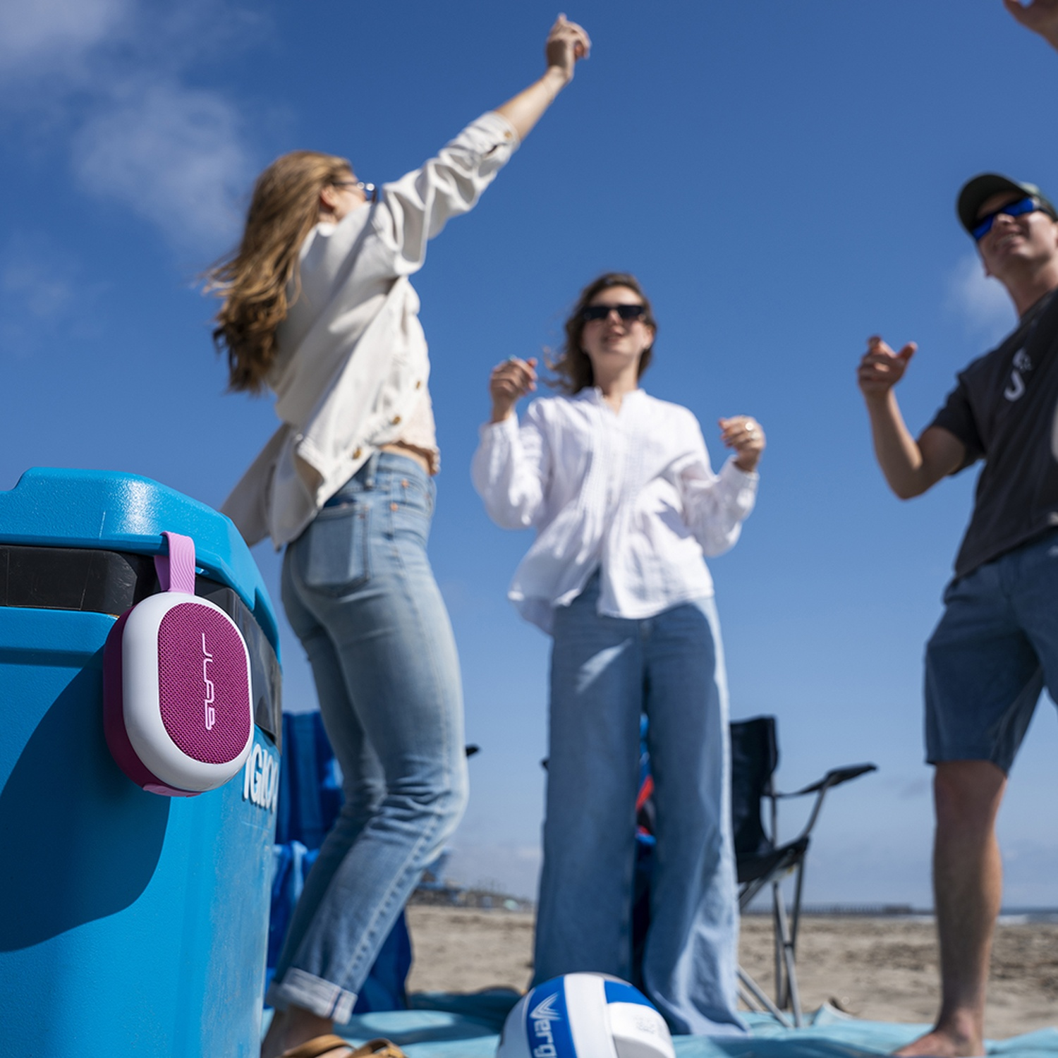 A purple speaker hangs on a blue cooler at the beach. Three people dance in the background.