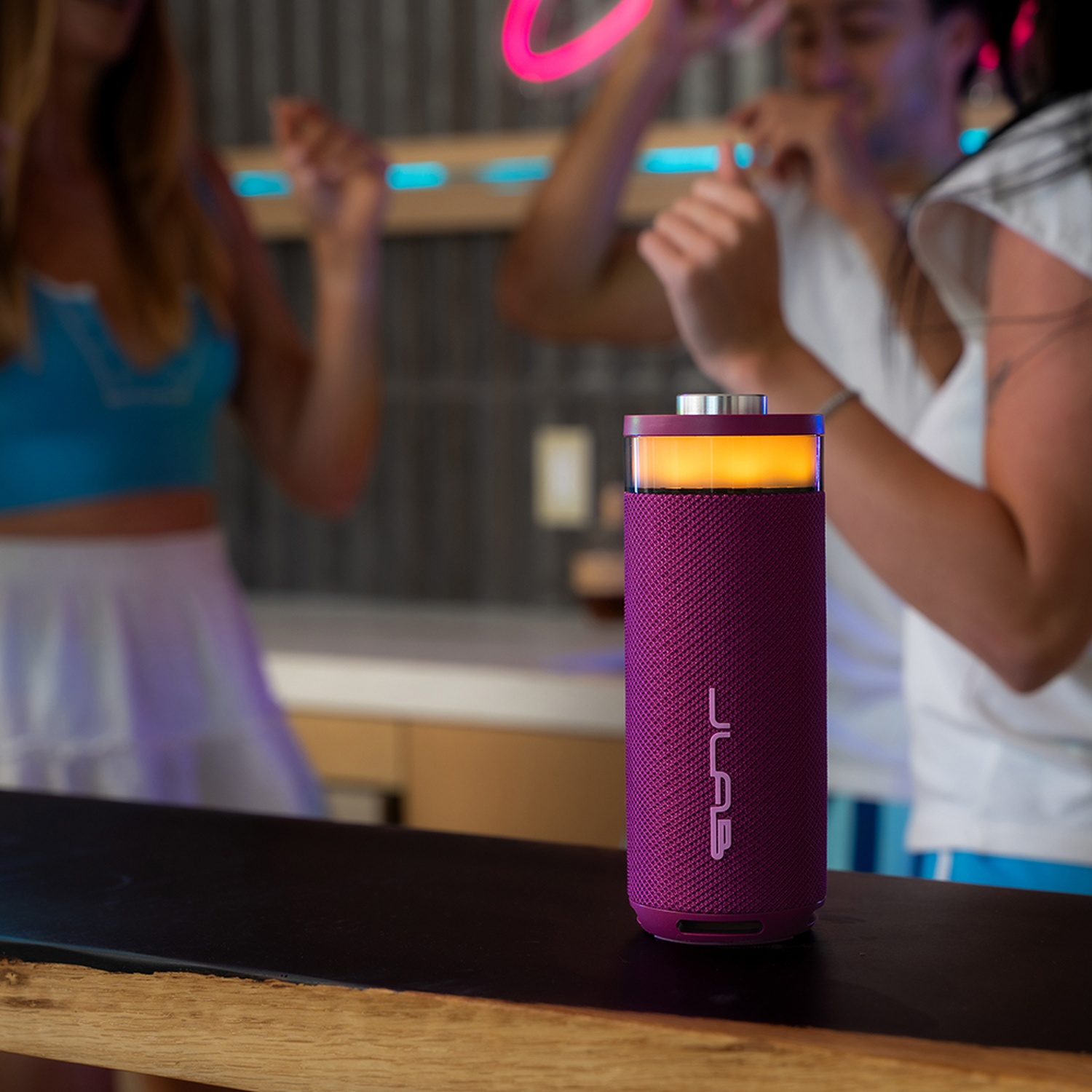 A purple speaker sits on a wooden counter with dancing people in neon light in the background.