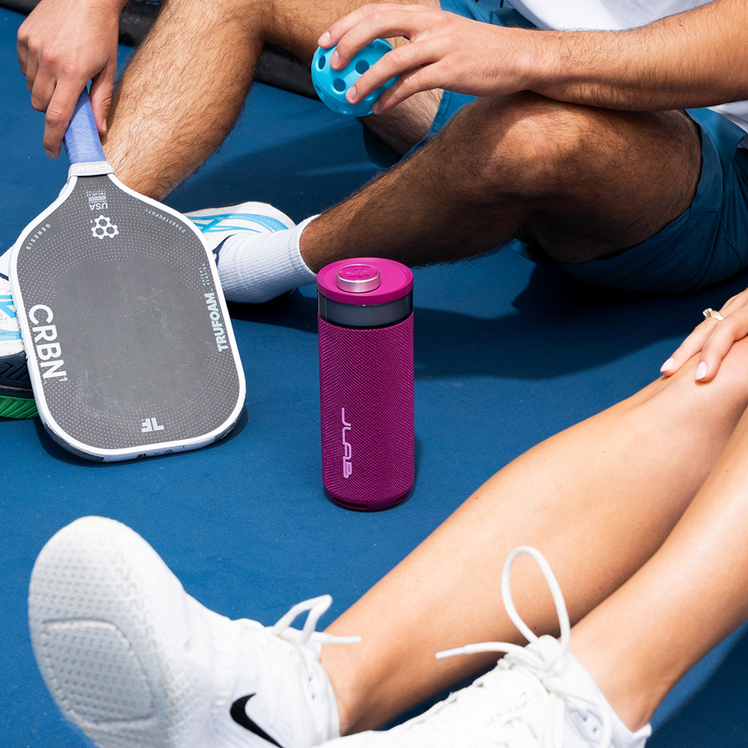 Two people sit on a blue court with a paddle, ball, and water bottle.
