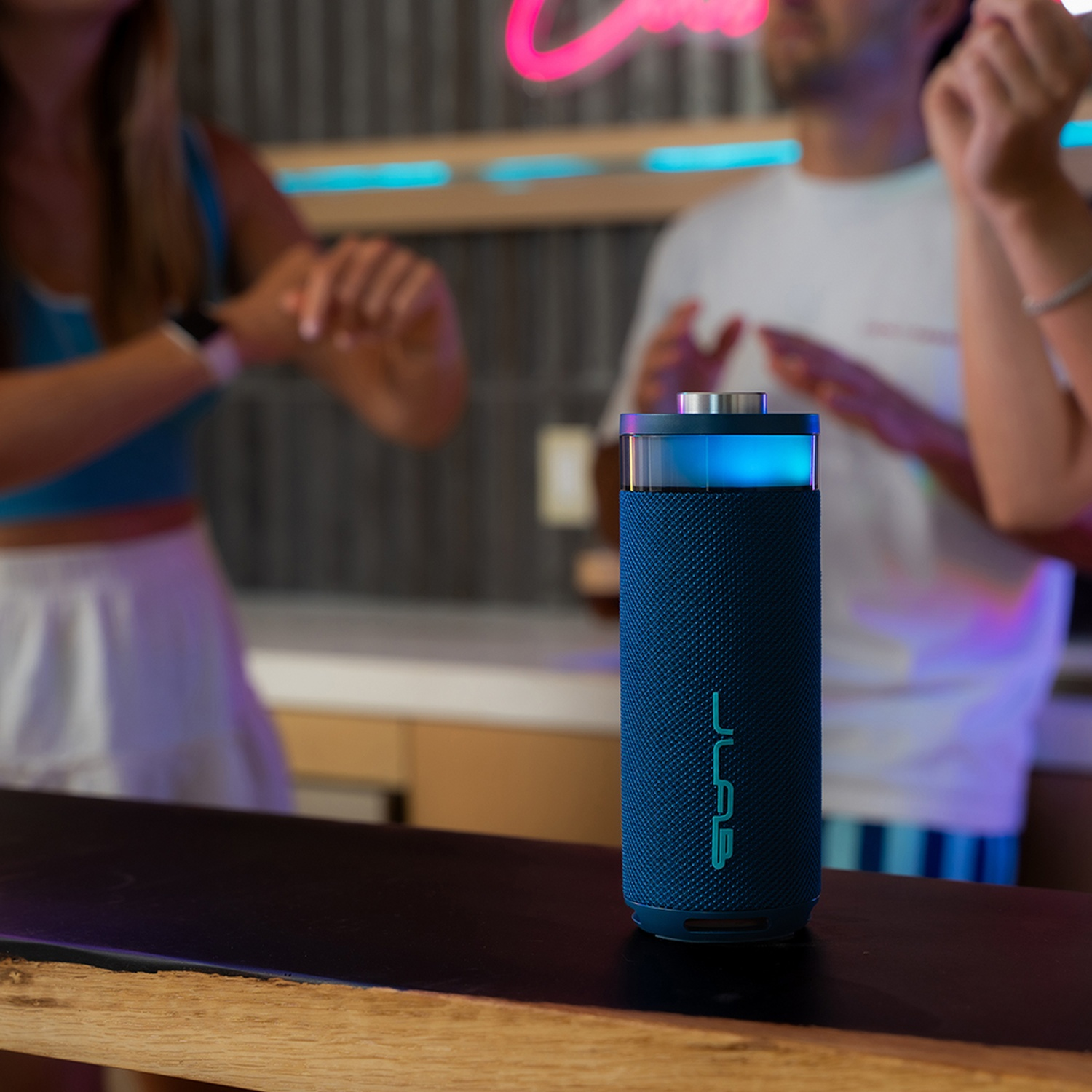 A blue speaker sits on a wooden counter with people dancing in the background.