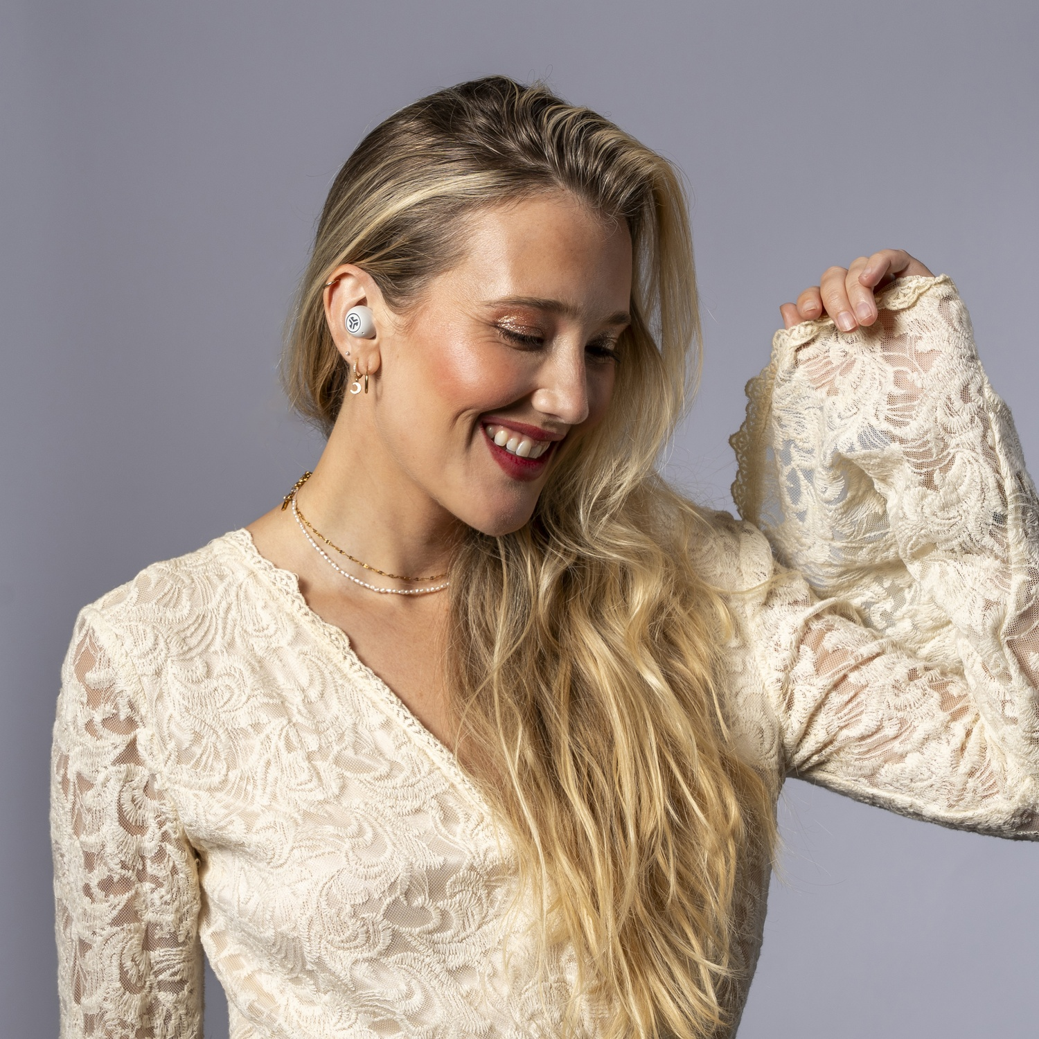 Een lachende vrouw met blond haar draagt een kanten jurk en witte oordopjes.