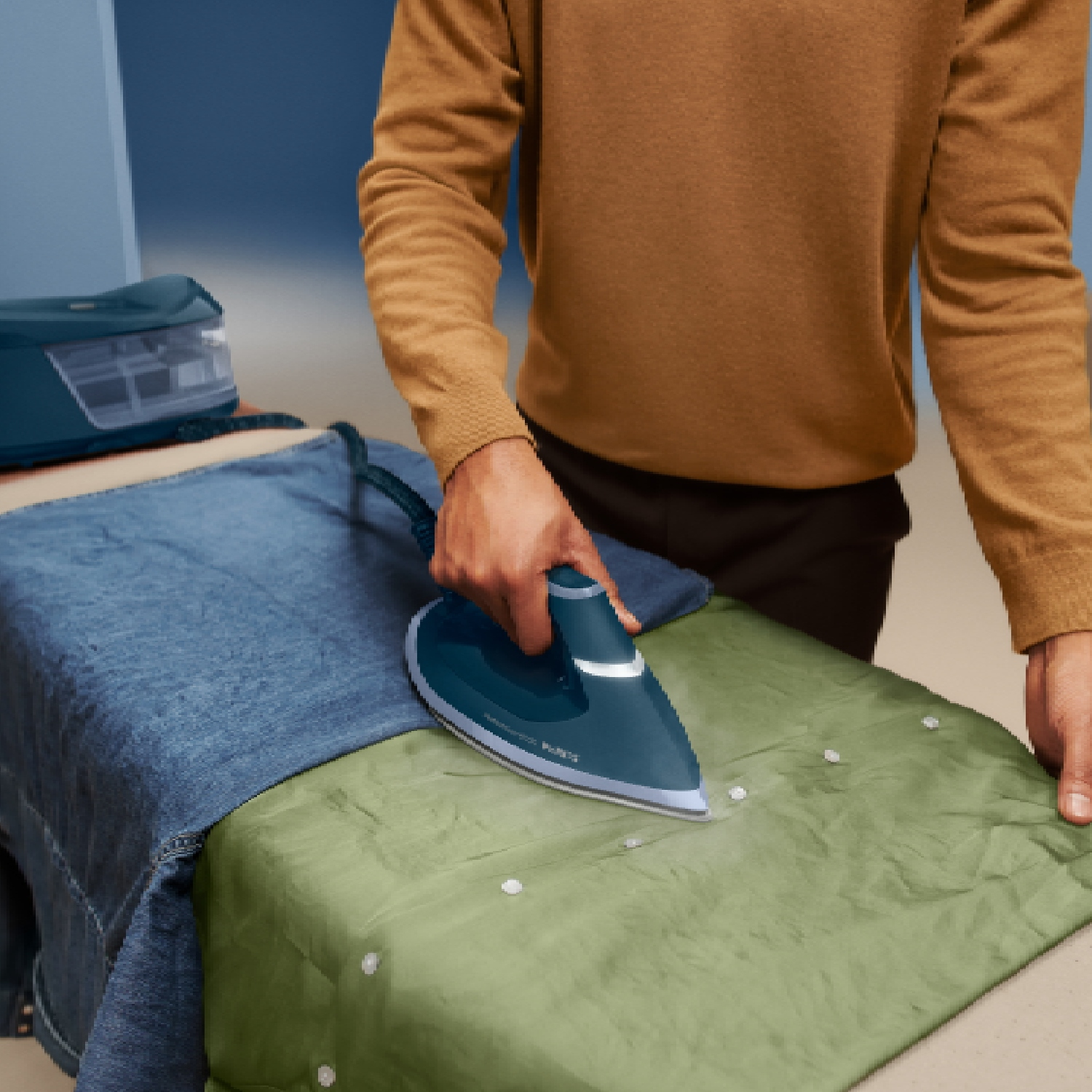 Persona planchando jeans azules y tela verde en una tabla de planchar con plancha de vapor.