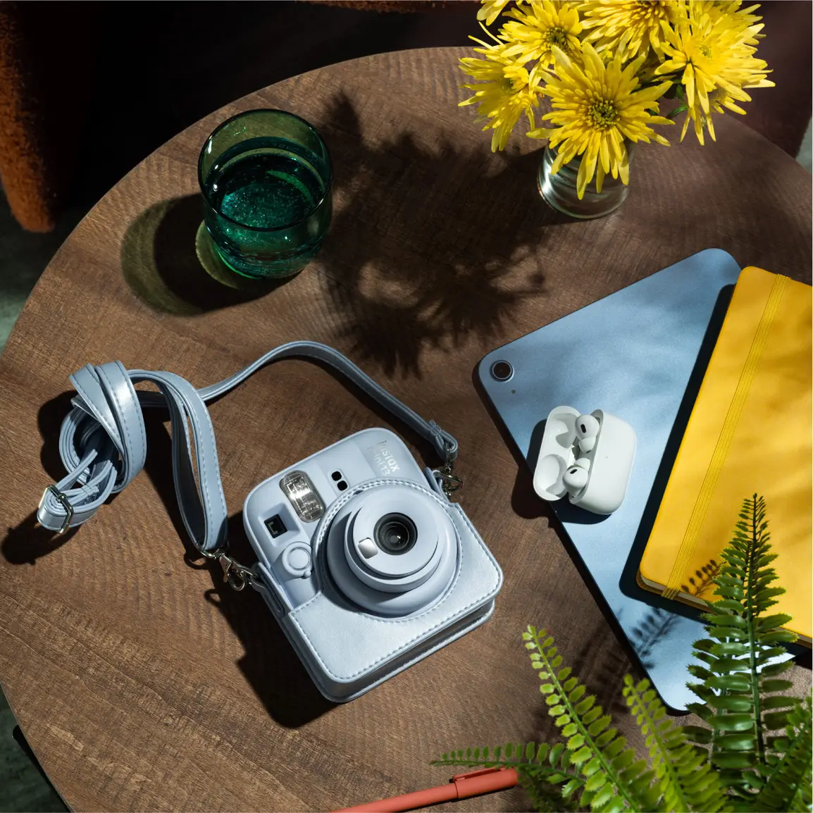 Une camera instantanee bleu clair avec sangle repose sur table en bois a cote d'un verre de liquide vert.