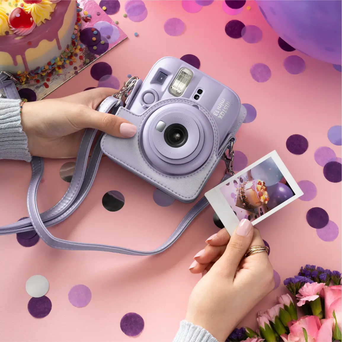 Mains tenant un appareil photo instantané violet et une photo sur fond rose avec confettis.