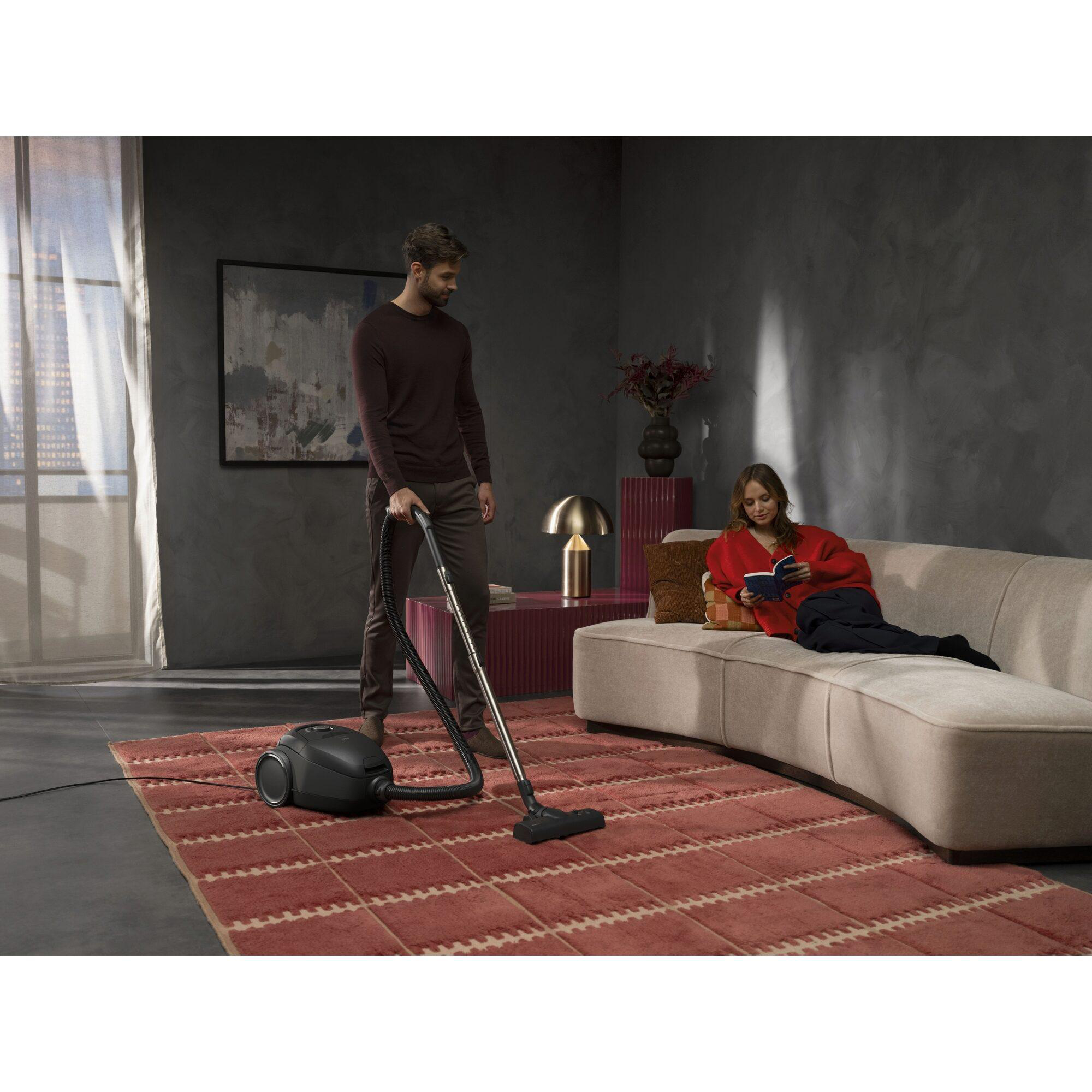 Mann saugt Teppich, Frau liest auf Sofa in modernem Wohnzimmer.